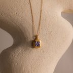 The Ribbed Alexandrite Pendant Necklace features a gold ribbed chain with a purple gemstone pendant, showcased on a textured, neutral-toned sculpted surface.