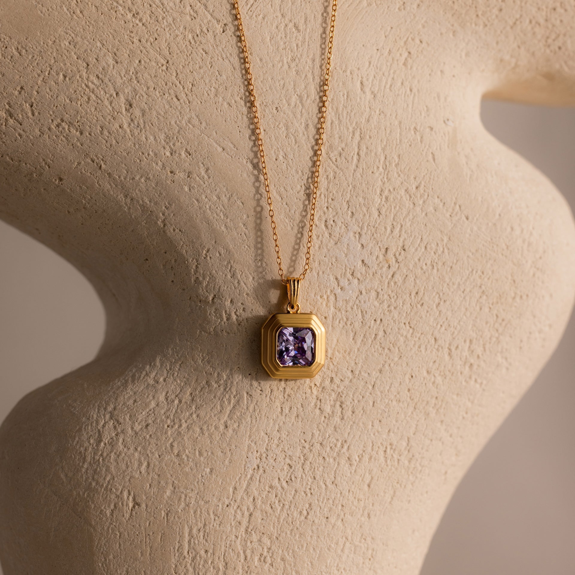 The Ribbed Alexandrite Pendant Necklace features a gold ribbed chain with a purple gemstone pendant, showcased on a textured, neutral-toned sculpted surface.