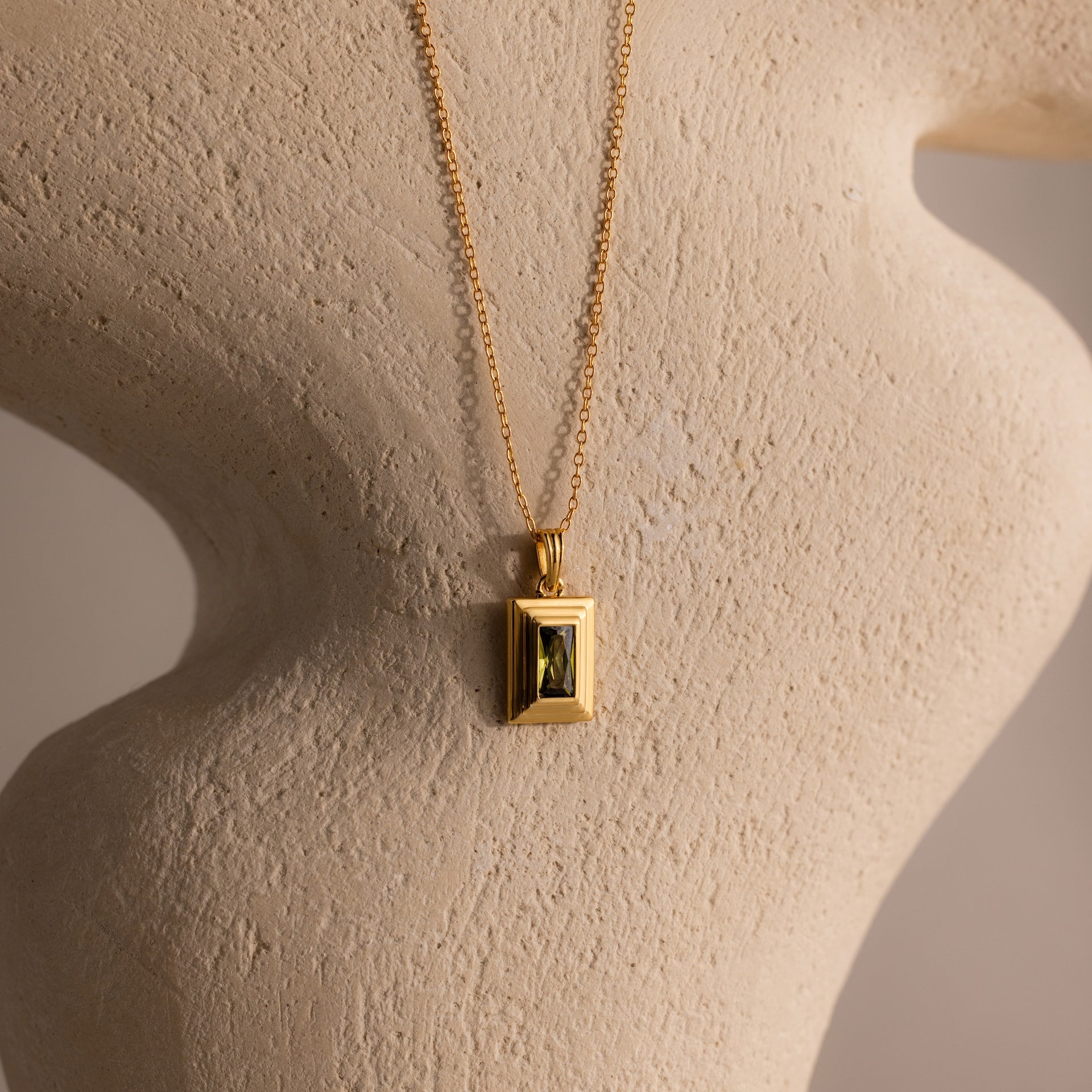 The Ribbed Peridot Pendant Necklace is shown with its rectangular pendant on a textured, light-colored sculpted surface.