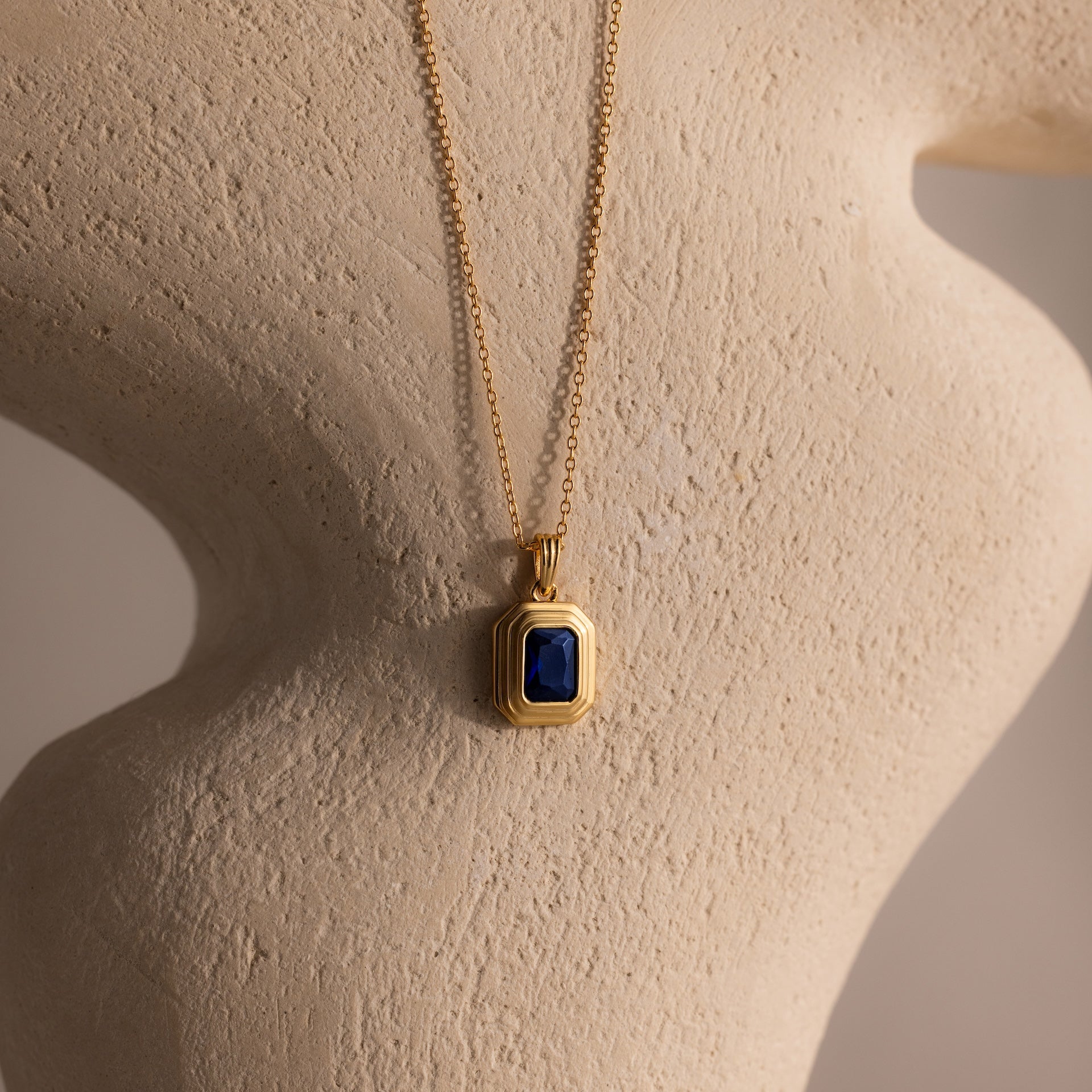 Ribbed Sapphire Pendant Necklace with a ribbed gold chain and a blue rectangular pendant, showcased on a textured beige sculpture.
