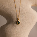 The Ribbed Emerald Pendant Necklace features a teardrop-shaped green gemstone displayed on a textured beige sculpture.