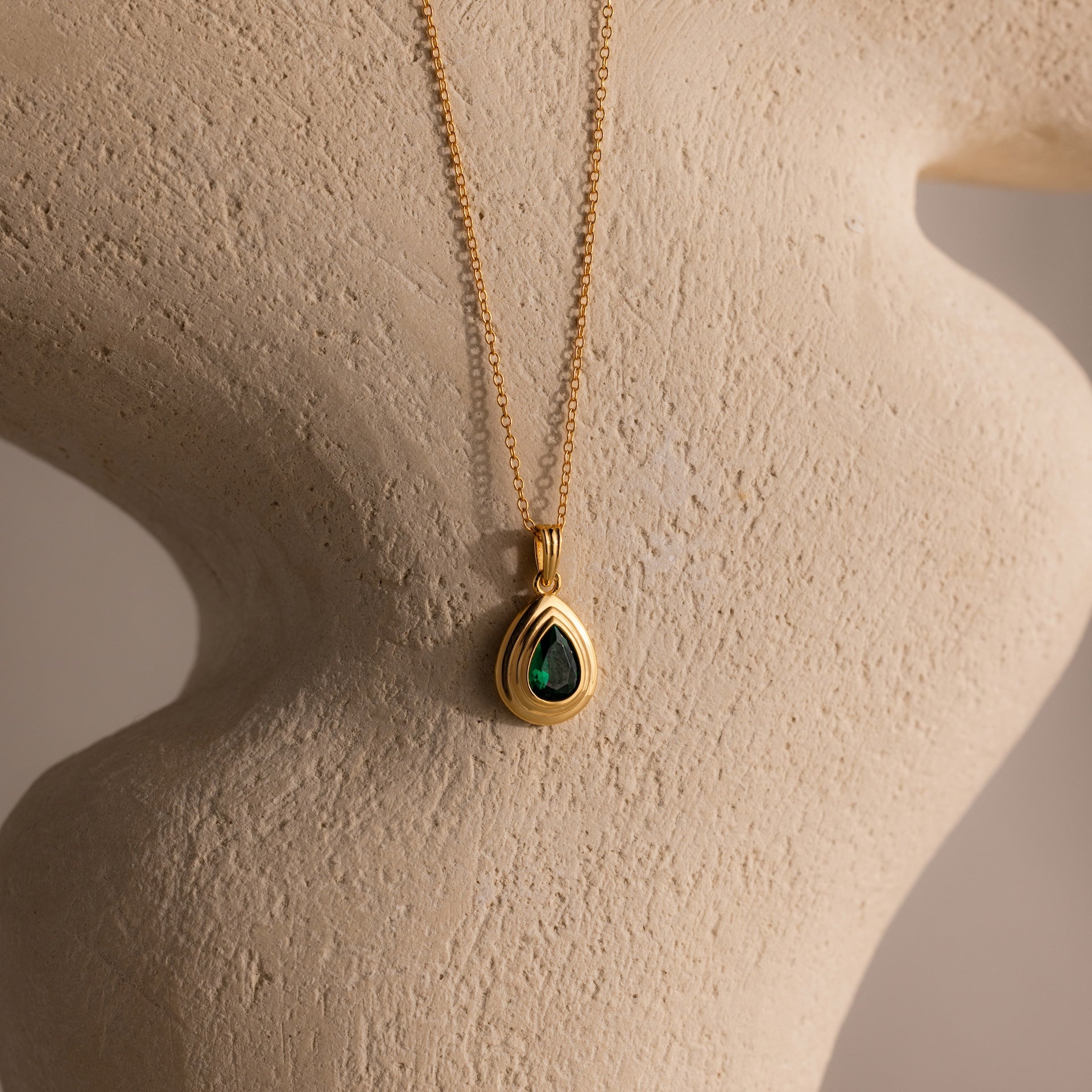 The Ribbed Emerald Pendant Necklace features a teardrop-shaped green gemstone displayed on a textured beige sculpture.