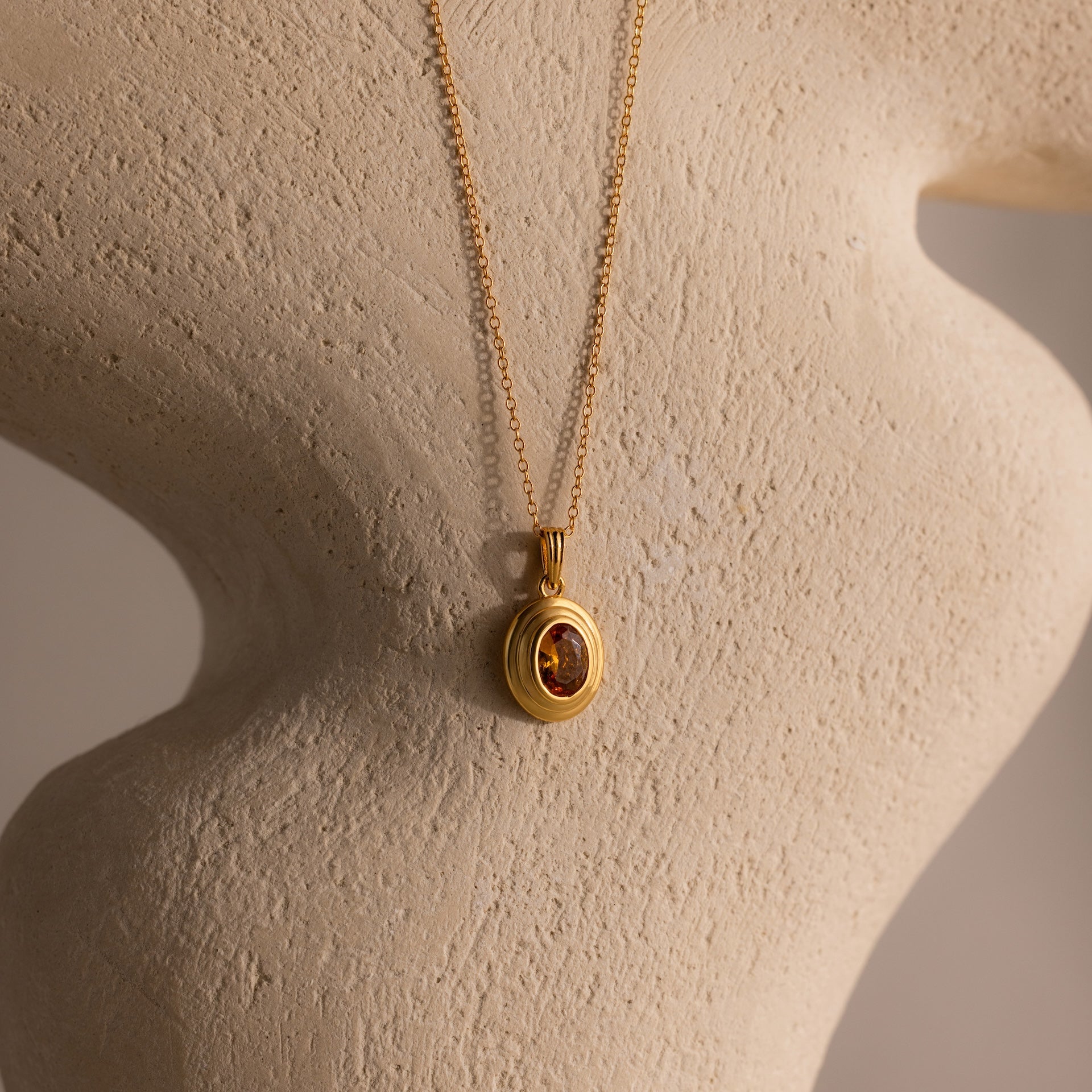 The Ribbed Topaz Pendant Necklace, featuring a ribbed gold chain with an oval red gemstone pendant, is elegantly displayed on a textured beige bust.