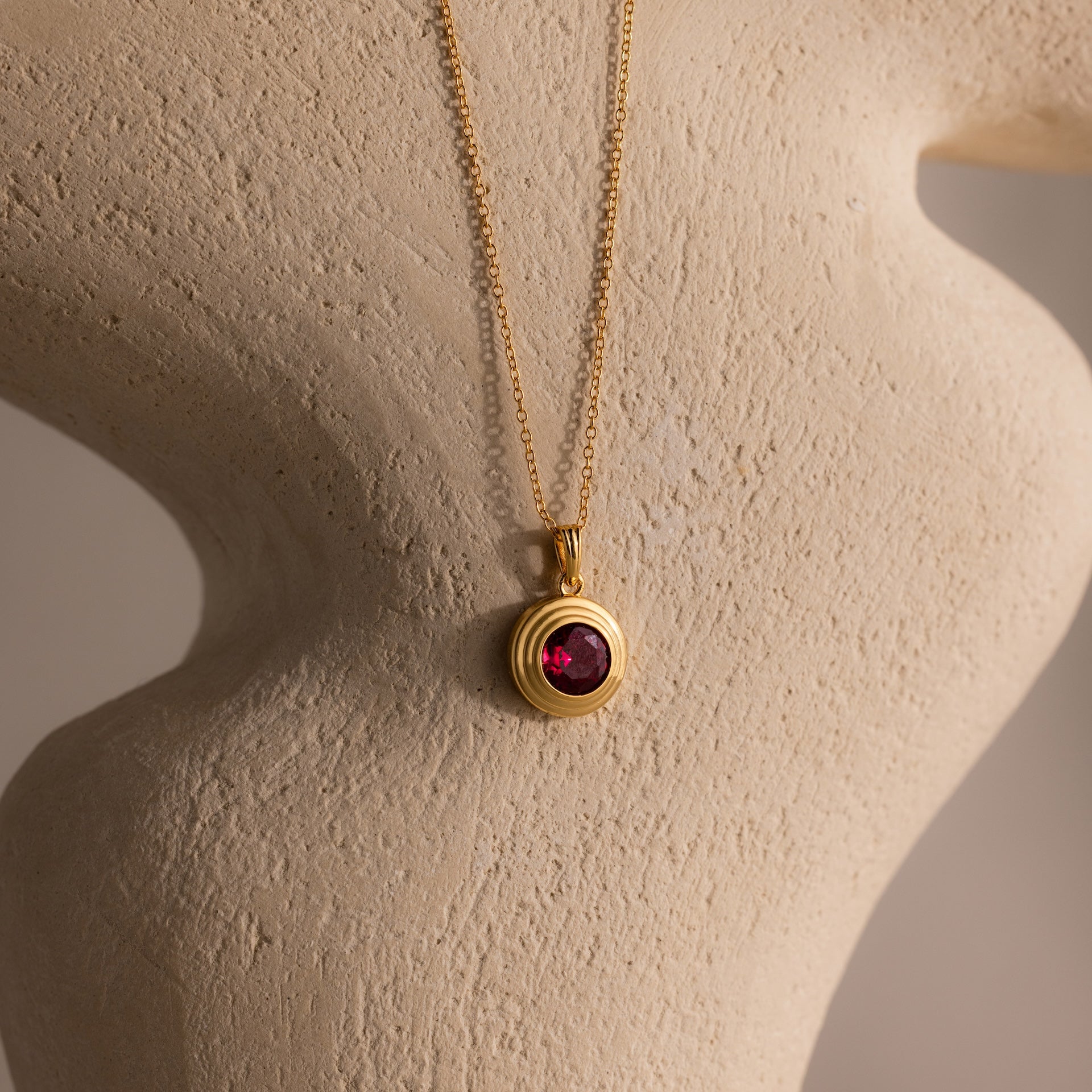 The Ribbed Ruby Pendant Necklace features a gold ribbed chain and round red gemstone pendant, ideal as a birthstone piece, displayed on a textured beige mannequin bust.