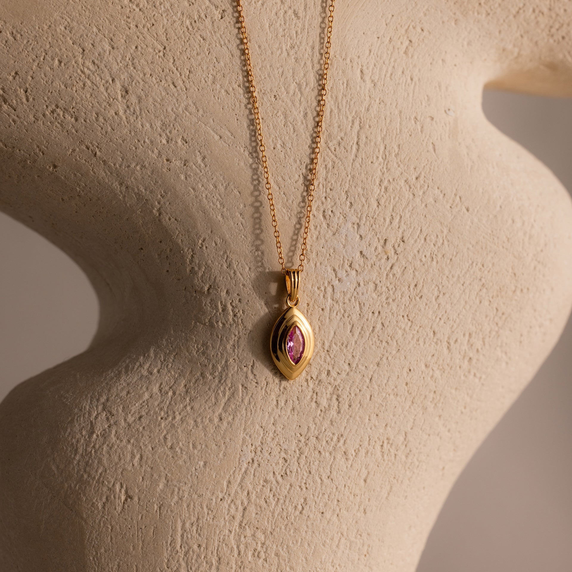 The Ribbed Pink Tourmaline Pendant Necklace is displayed on a textured beige sculpture background.