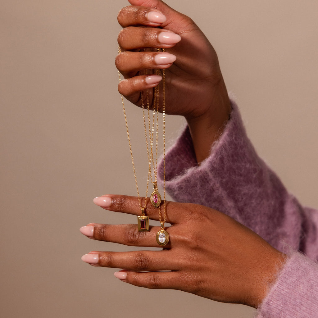 A hand holds three gold gemstone necklaces, including the Ribbed Garnet Pendant Necklace and a colorful birthstone pendant necklace, against a beige background.