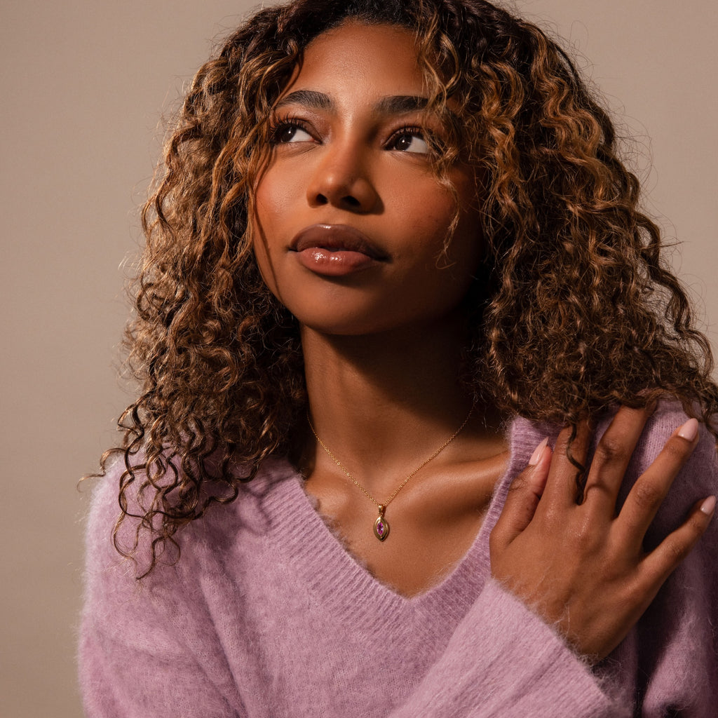 Woman with curly hair in a fuzzy lavender sweater looks upward, hand on shoulder, wearing the Ribbed Pink Tourmaline Pendant Necklace that catches the light.