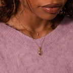 A woman in a lavender sweater wears a Ribbed Amethyst Pendant Necklace, featuring a ribbed gold chain with a purple amethyst pendant.