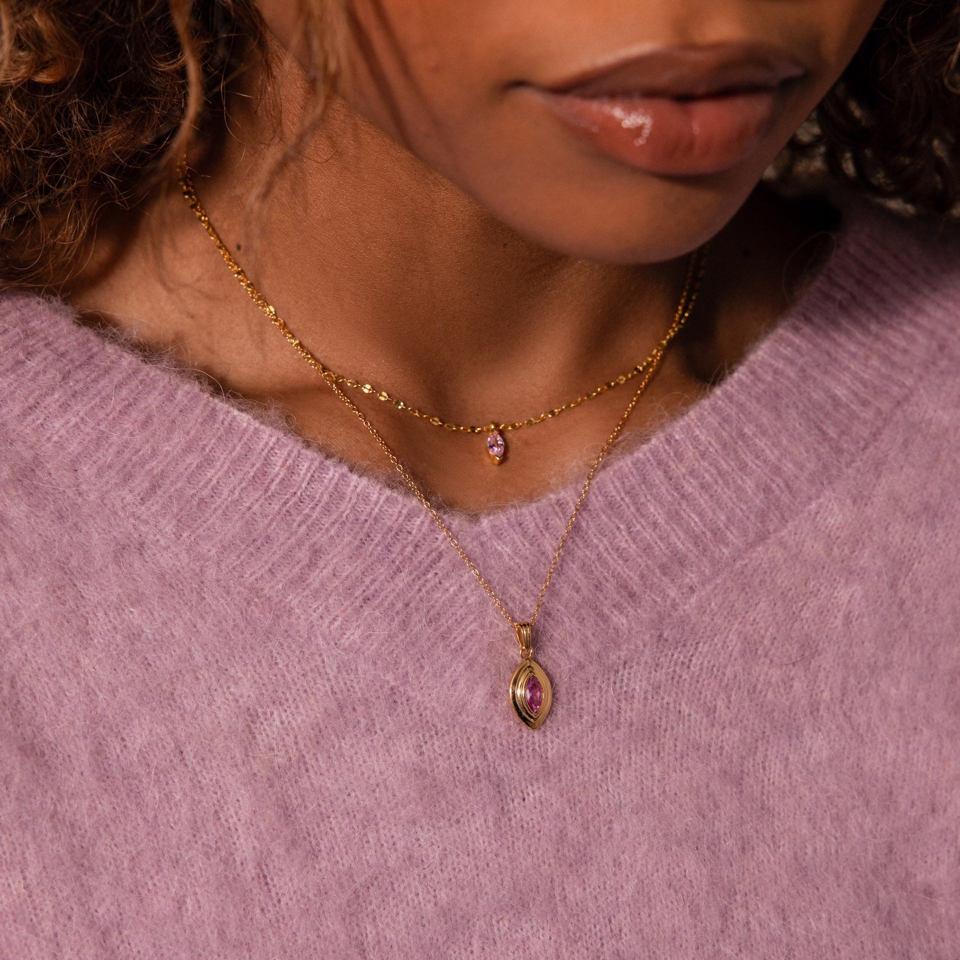 A woman in a lavender sweater wears a Ribbed Amethyst Pendant Necklace, featuring a ribbed gold chain with a purple amethyst pendant.