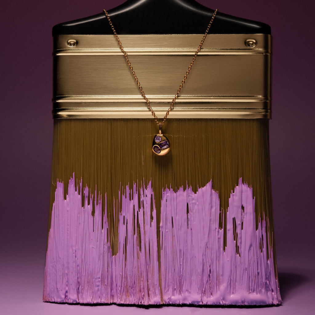 A gold paintbrush with purple paint is displayed, featuring the Mixed Amethyst Capsule Pendant Necklace draped elegantly over its handle.