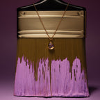 A gold paintbrush with purple paint is displayed, featuring the Mixed Amethyst Capsule Pendant Necklace draped elegantly over its handle.