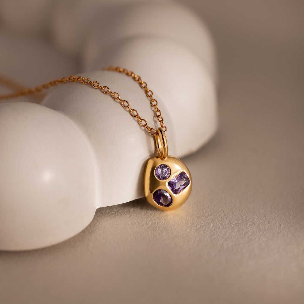 Mixed Amethyst Capsule Pendant Necklace featuring three purple gemstones, displayed on a smooth white surface under soft, warm lighting.