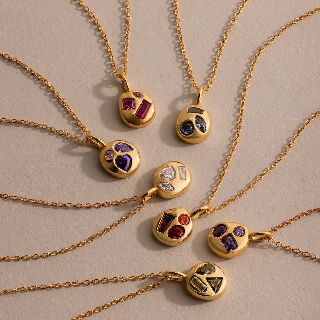 Seven gold necklaces, including the Mixed Amethyst Capsule Pendant Necklace plus birthstone and capsule styles, arranged in a vibrant circular pattern.