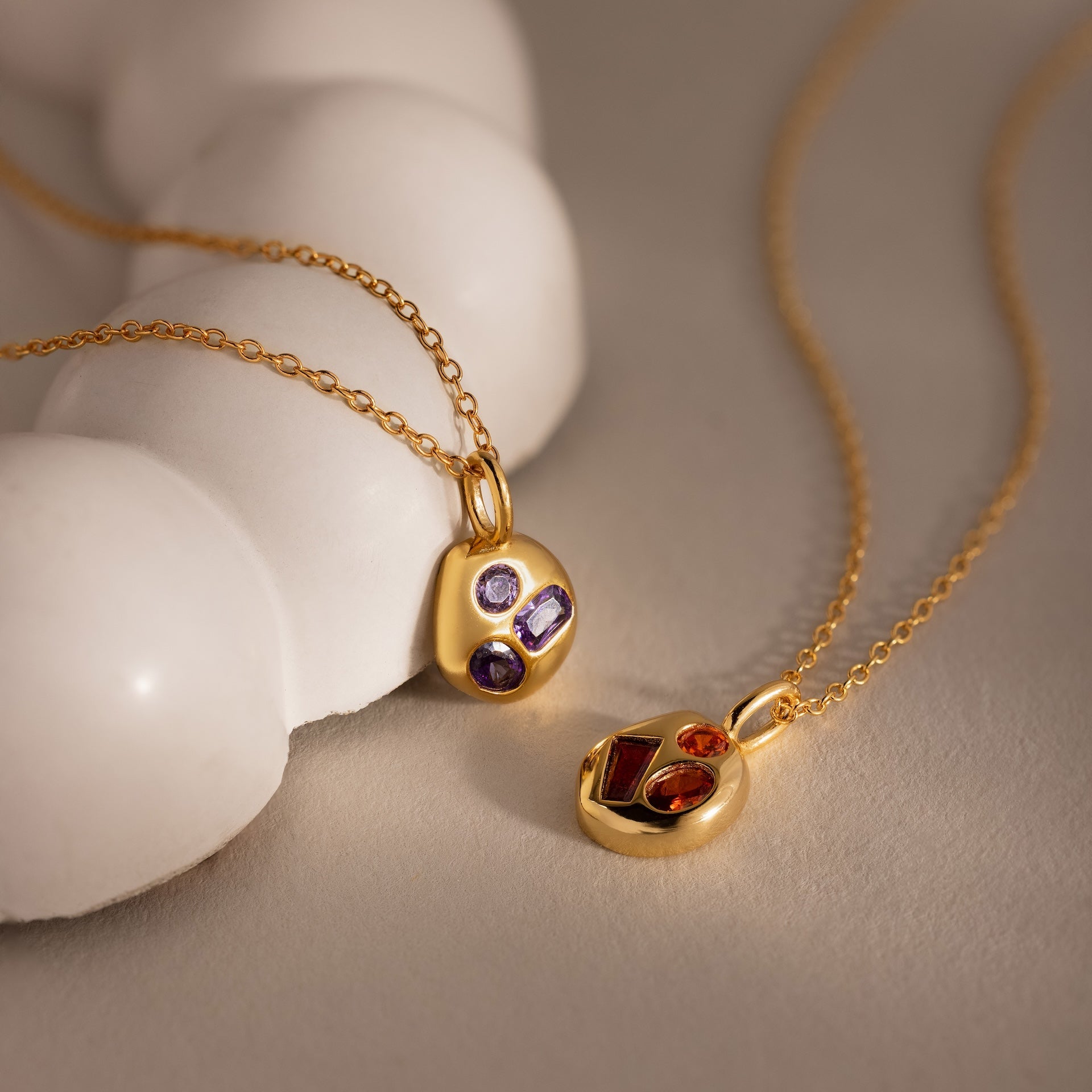 Two gold Mixed Birthstone Capsule Necklaces, one with purple stones and one with red, rest on a soft beige surface—an ideal set for a meaningful touch.