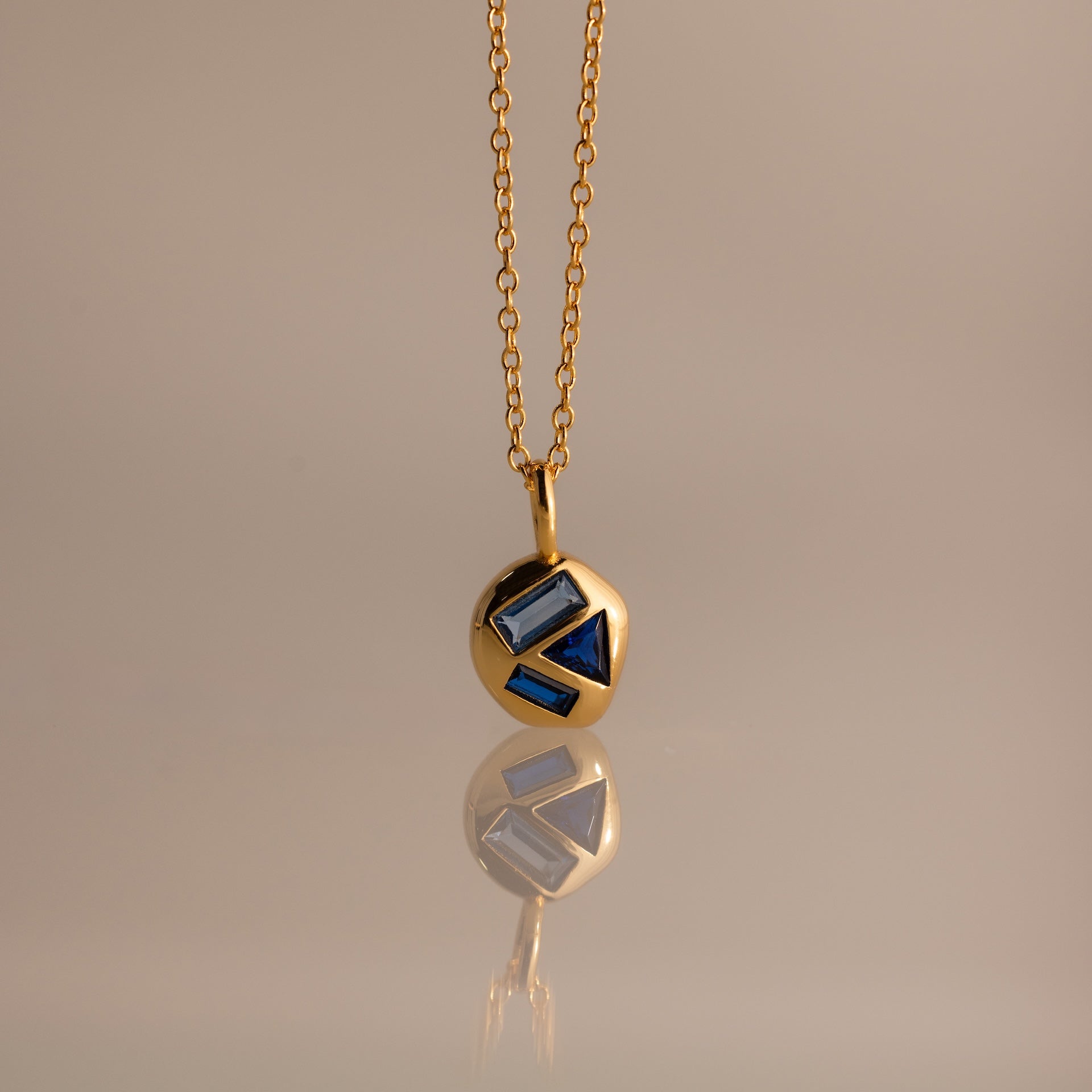 Mixed Sapphire Capsule Pendant Necklace featuring a gold round capsule pendant with geometric blue sapphire inlays, displayed on a neutral background.