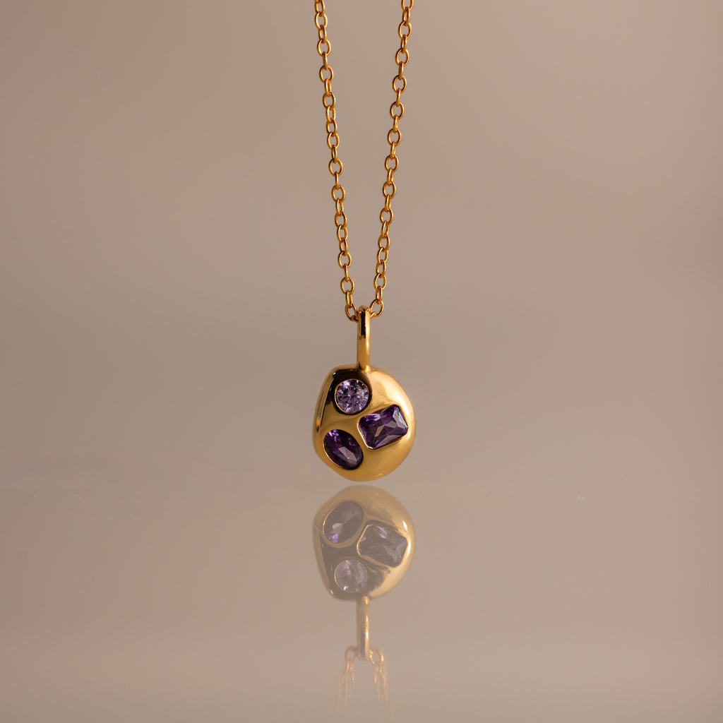 The Mixed Amethyst Capsule Pendant Necklace features a gold capsule design with a round pendant and three small purple gemstones, beautifully reflected on a neutral surface.