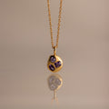 The Mixed Amethyst Capsule Pendant Necklace features a gold capsule design with a round pendant and three small purple gemstones, beautifully reflected on a neutral surface.