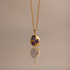 The Mixed Amethyst Capsule Pendant Necklace features a gold capsule design with a round pendant and three small purple gemstones, beautifully reflected on a neutral surface.