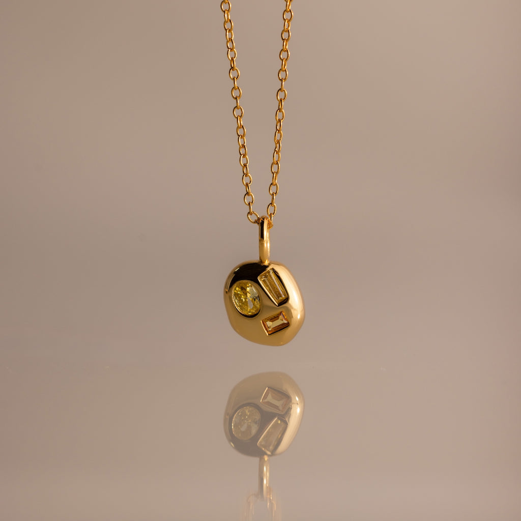 The Mixed Topaz Capsule Pendant Necklace features a gold design with three embedded topaz stones on a round charm—a timeless birthstone piece for personalizing your style, shown against a neutral background.