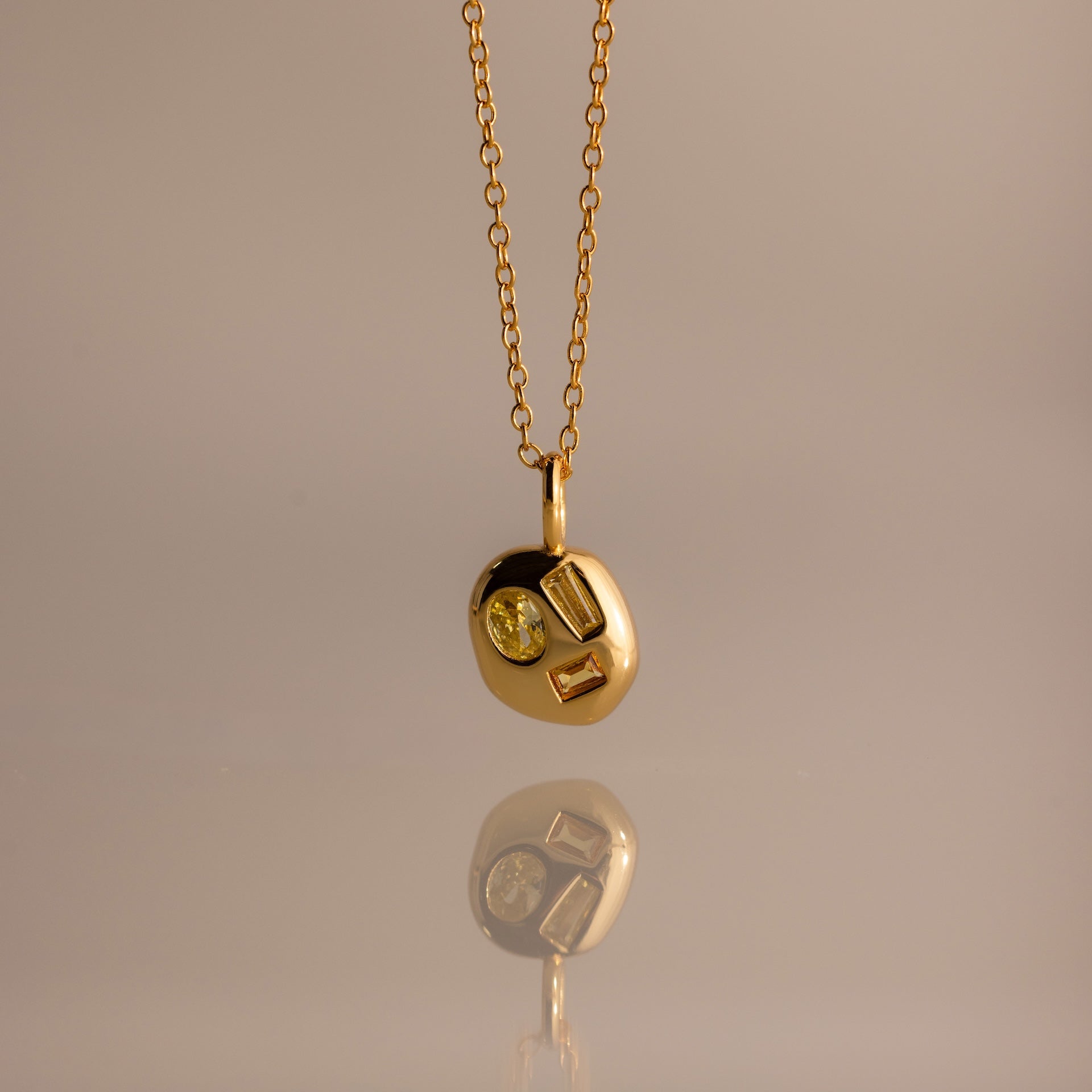 The Mixed Topaz Capsule Pendant Necklace features a gold design with three embedded topaz stones on a round charm—a timeless birthstone piece for personalizing your style, shown against a neutral background.