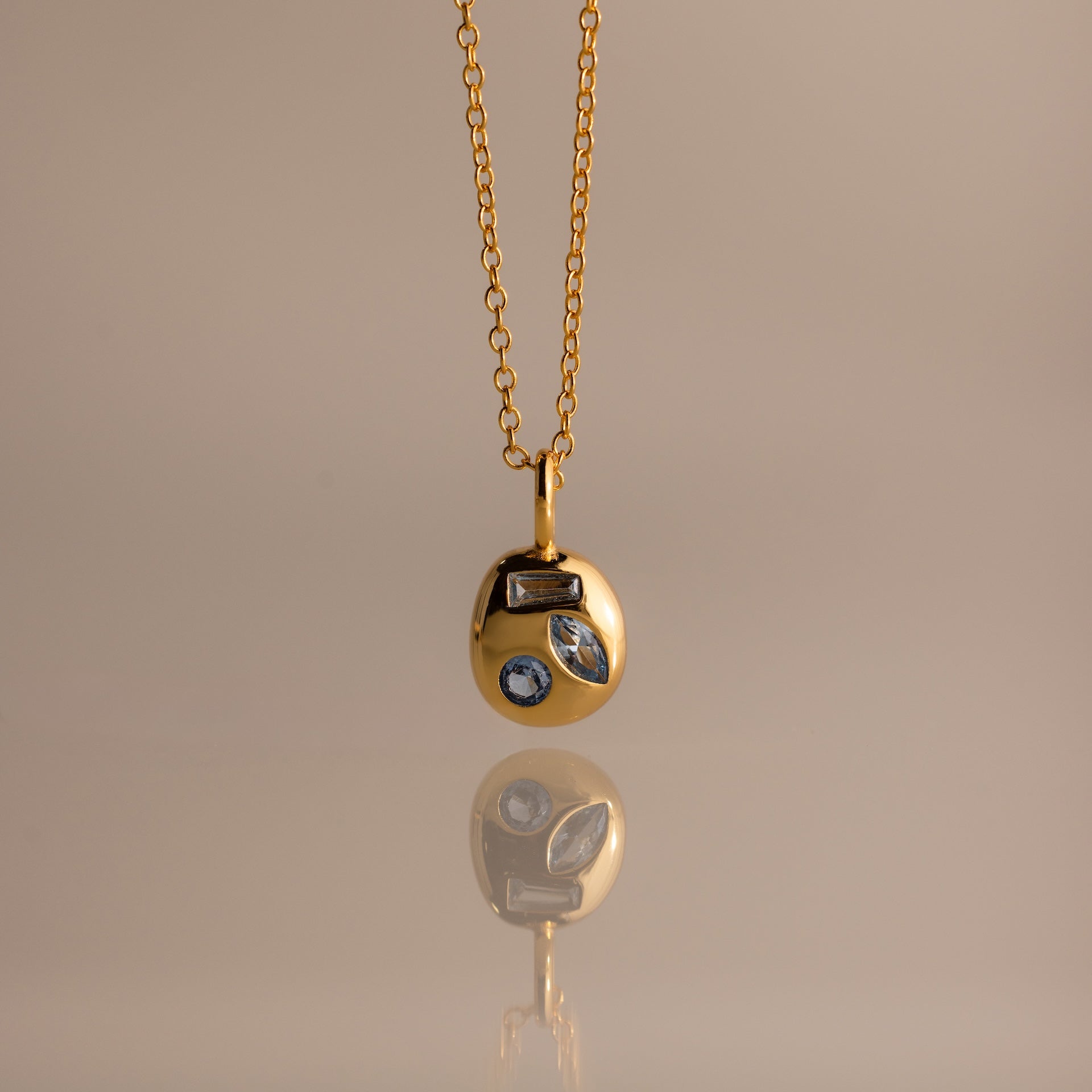 Mixed Aquamarine Capsule Pendant Necklace featuring a round charm with three different shapes of aquamarine gemstones, displayed on a beige background.