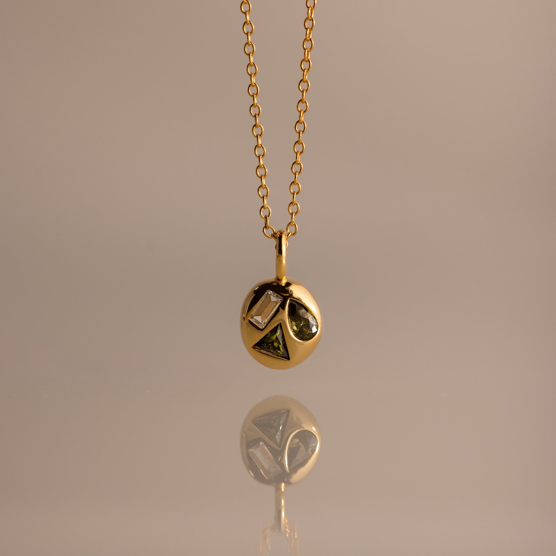 Mixed Peridot Capsule Pendant Necklace with a round pendant featuring geometric shapes, reflected on a smooth surface.