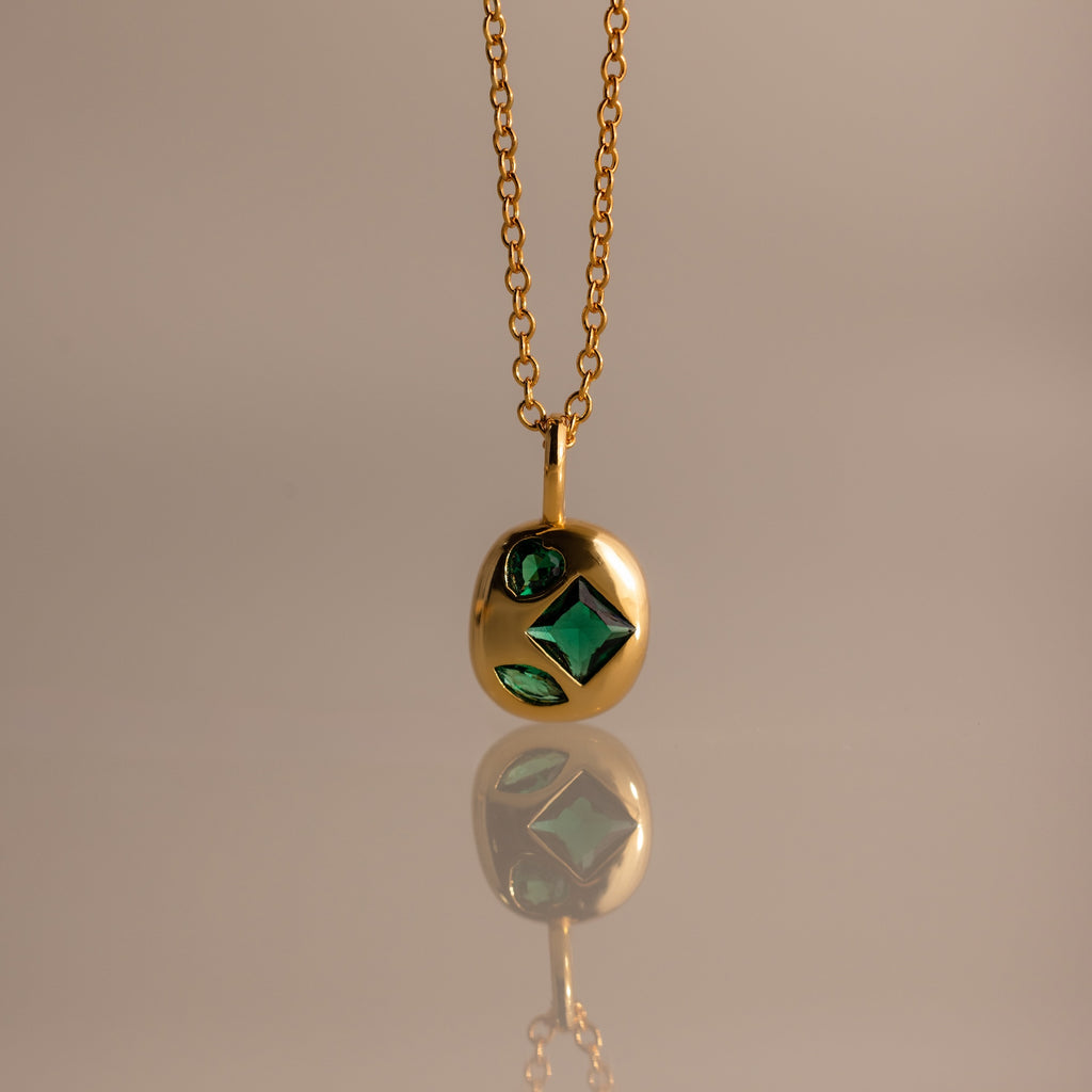Mixed Emerald Capsule Pendant Necklace featuring green gemstones in a capsule-shaped pendant, elegantly reflected on a smooth surface.