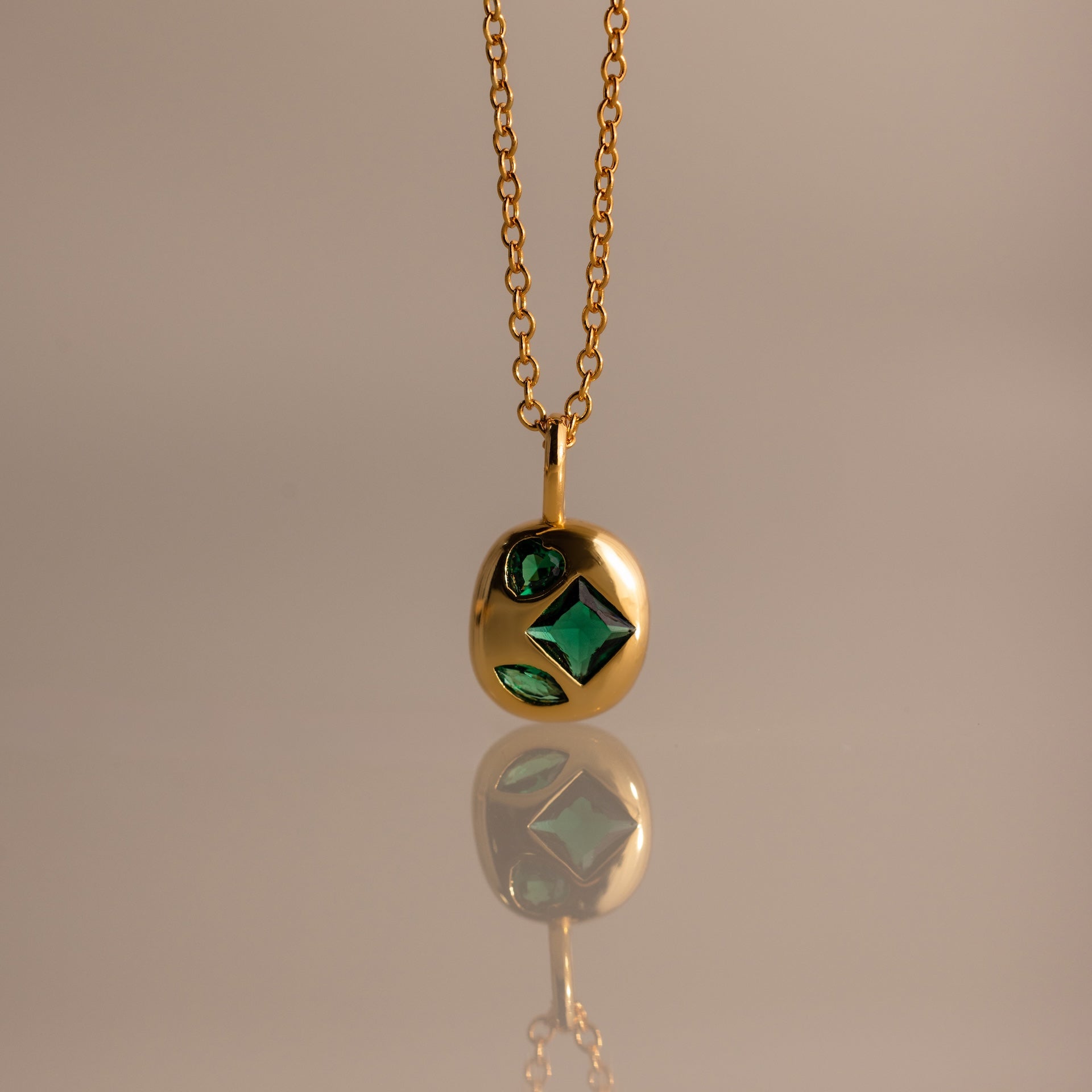 Mixed Emerald Capsule Pendant Necklace featuring green gemstones in a capsule-shaped pendant, elegantly reflected on a smooth surface.