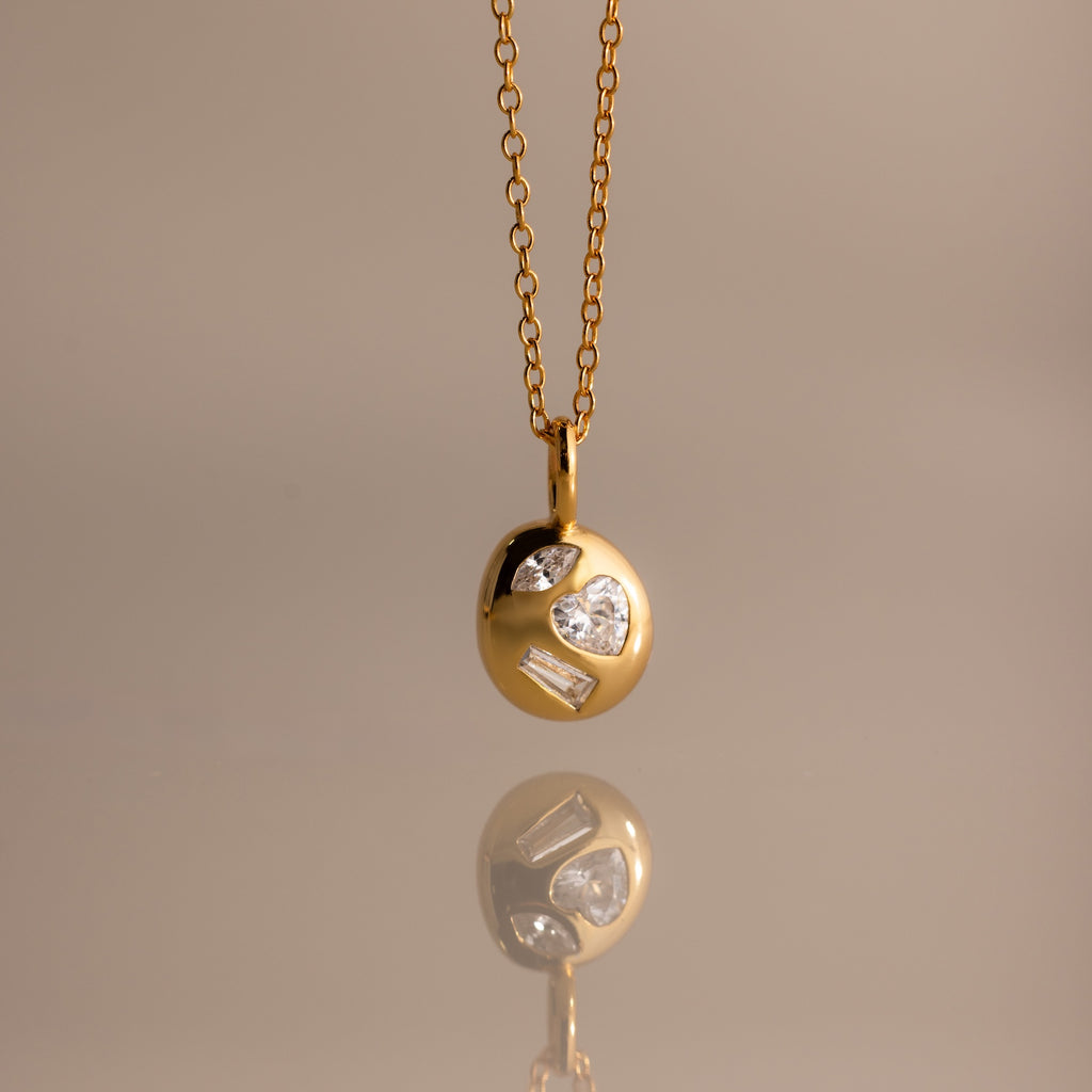 The Mixed Diamond Capsule Pendant Necklace features a gold capsule design with a round pendant set with clear gemstones, displayed on a neutral background.