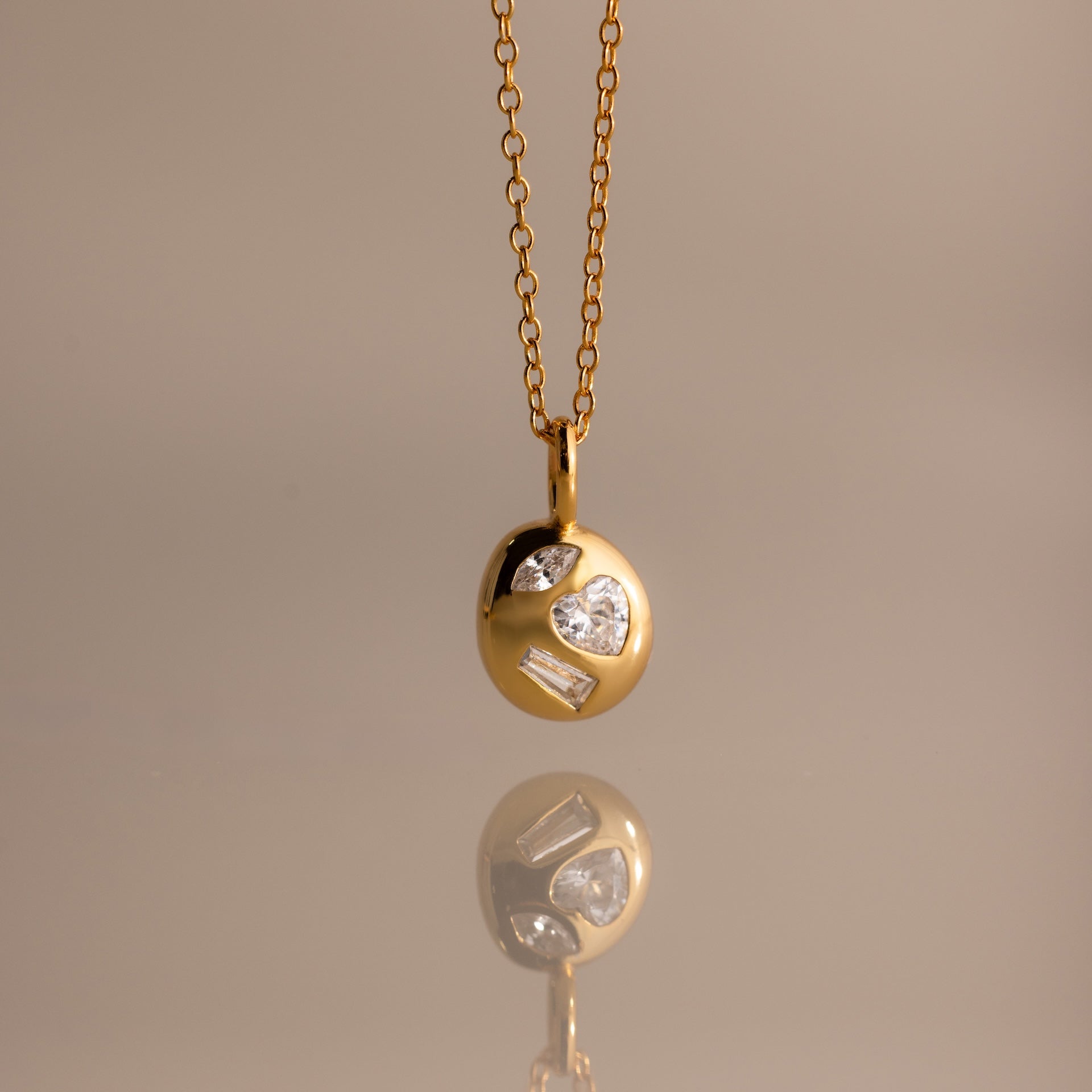 The Mixed Diamond Capsule Pendant Necklace features a gold capsule design with a round pendant set with clear gemstones, displayed on a neutral background.