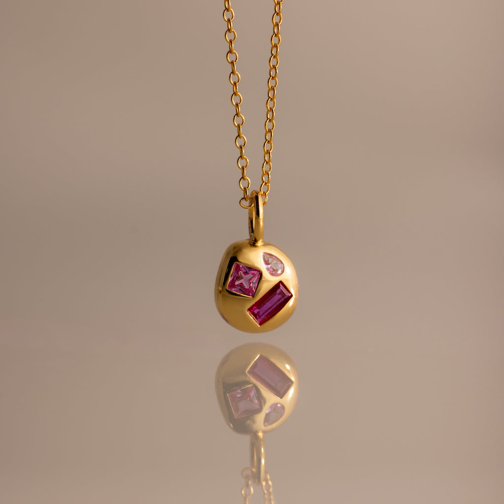 The Mixed Ruby Capsule Pendant Necklace—a round design with three pink and clear gemstones—rests elegantly reflected on a smooth surface.