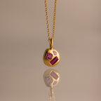 The Mixed Ruby Capsule Pendant Necklace—a round design with three pink and clear gemstones—rests elegantly reflected on a smooth surface.