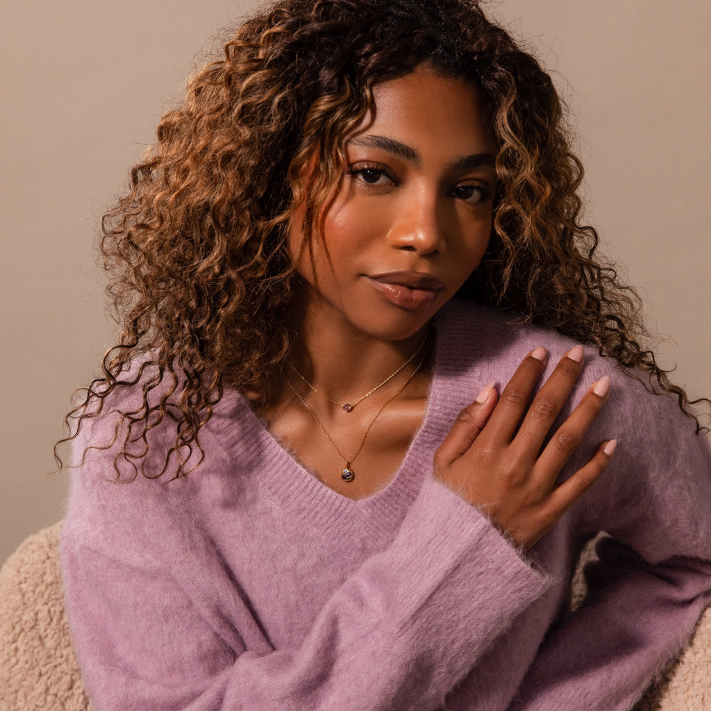 Woman with curly hair wearing a lavender sweater and the Mixed Amethyst Capsule Pendant Necklace, layered with other necklaces, sitting on a beige chair.