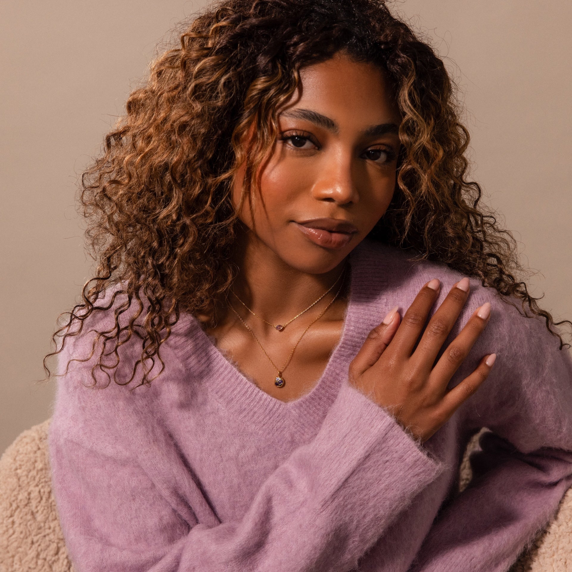 Woman with curly hair wearing a lavender sweater and the Mixed Amethyst Capsule Pendant Necklace, layered with other necklaces, sitting on a beige chair.