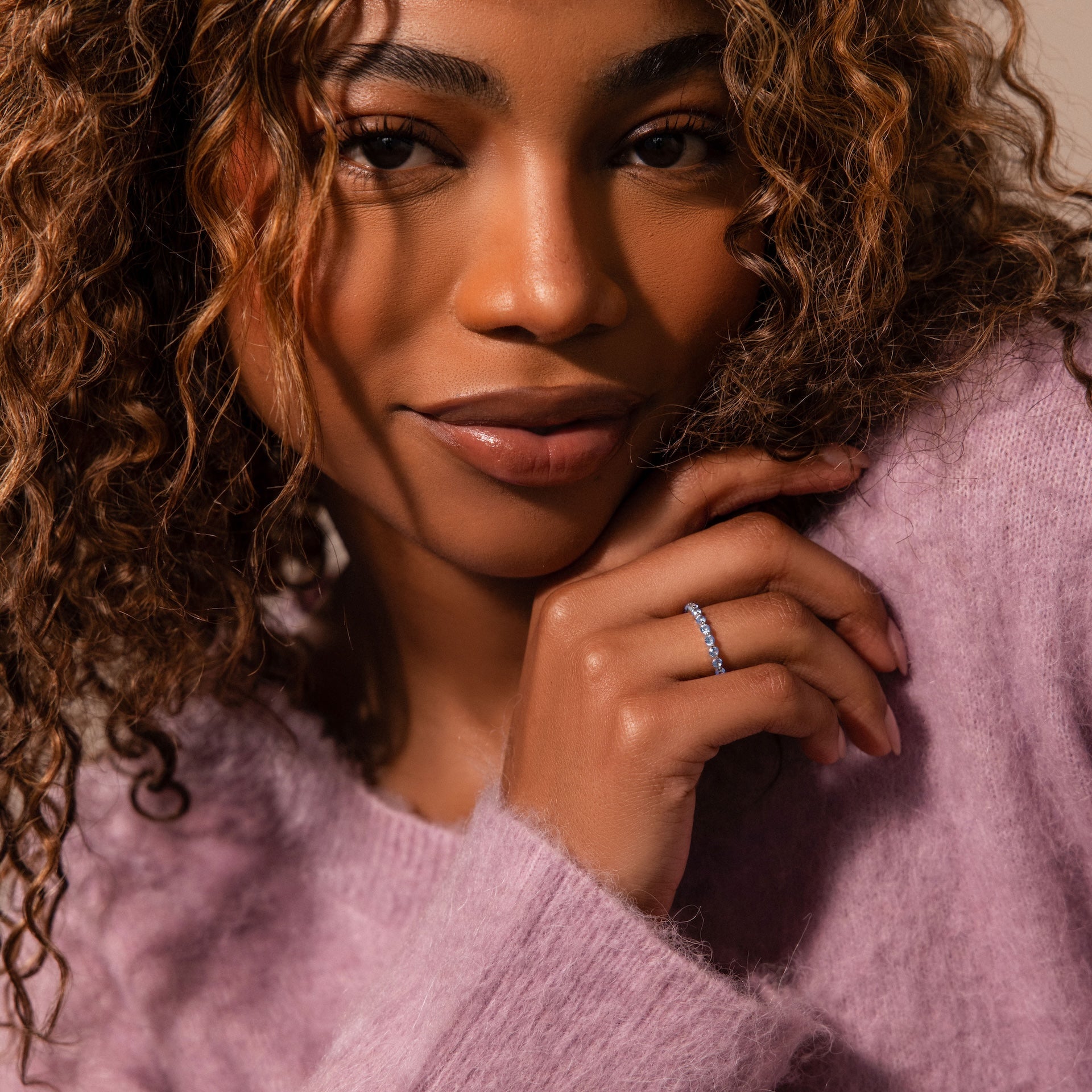 Woman with curly hair in a soft purple sweater smiles as she displays the sparkling Lace Eternity Aquamarine Ring on her finger.