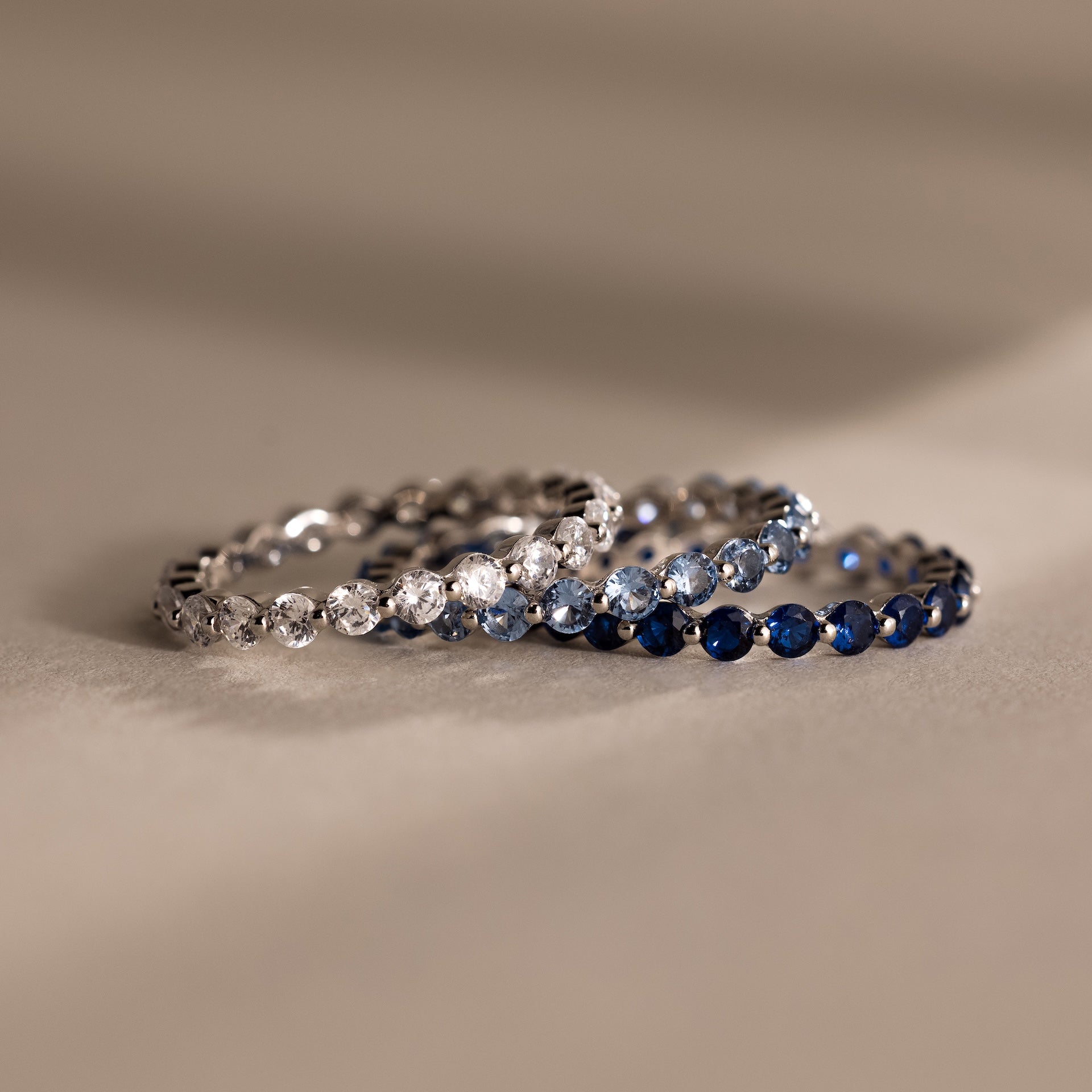 Two rings are placed side by side on a neutral surface: the Lace Eternity Blue Zircon Ring with sparkling blue stones, and an elegant eternity ring set with clear stones.