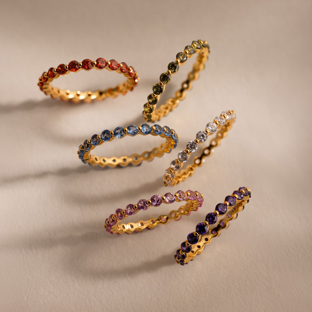 Six gold bands, including a shimmering Lace Eternity Diamond Ring, with sparkling gemstones are arranged in a neat arc on a beige background.