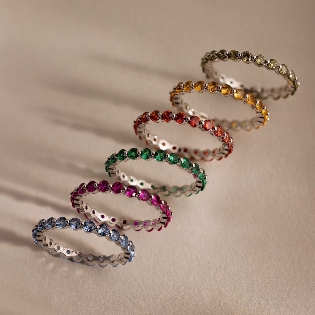 Six gemstone rings, featuring the Lace Eternity Emerald Ring, are arranged diagonally on a light beige surface with soft shadows.