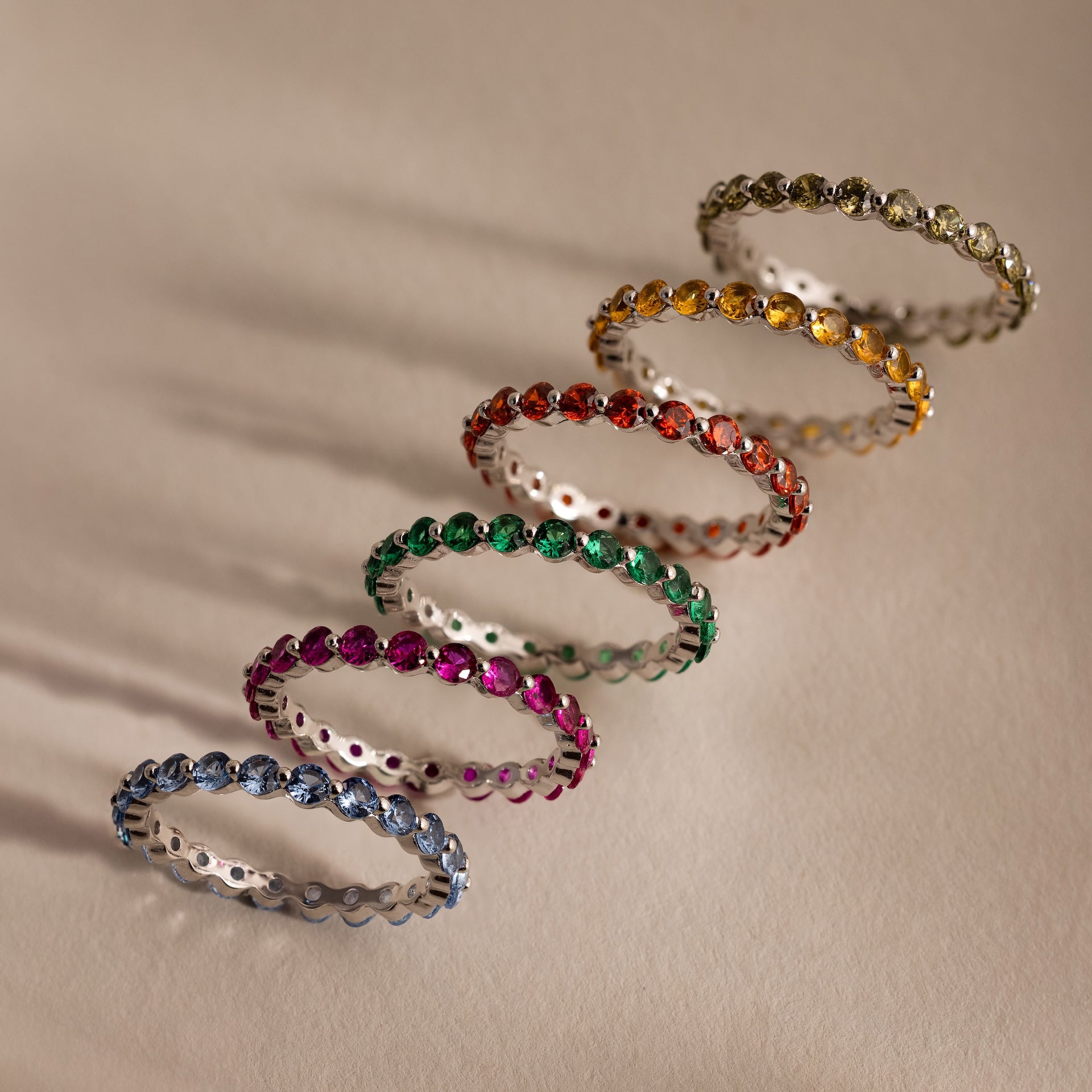 Six gemstone rings, featuring the Lace Eternity Emerald Ring, are arranged diagonally on a light beige surface with soft shadows.