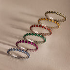Six Lace Eternity Topaz Rings in vibrant blue, pink, green, red, orange, and yellow are arranged in a diagonal line on a beige surface.