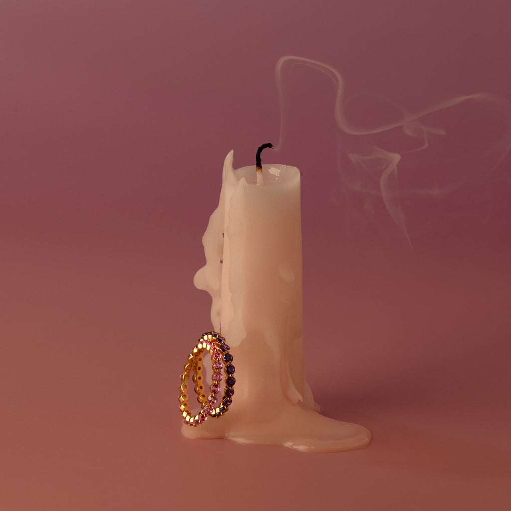A melted candle with smoke rising and two gold rings, including the Lace Eternity Birthstone Ring, leaning against its base on a pink background.