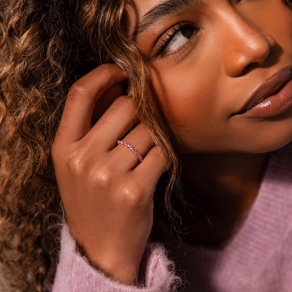 A woman with curly hair touches her ear, wearing the Lace Eternity Pink Tourmaline Ring and a soft pink sweater adorned with delicate lace accents.