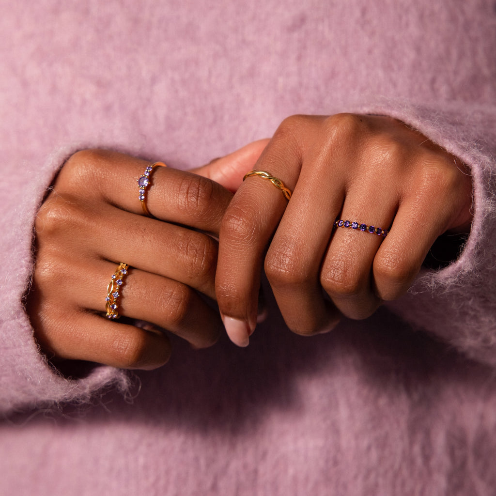 Two hands display gold and gemstone rings, featuring the Lace Eternity Pink Tourmaline Ring, with a light pink fuzzy sweater in the background.