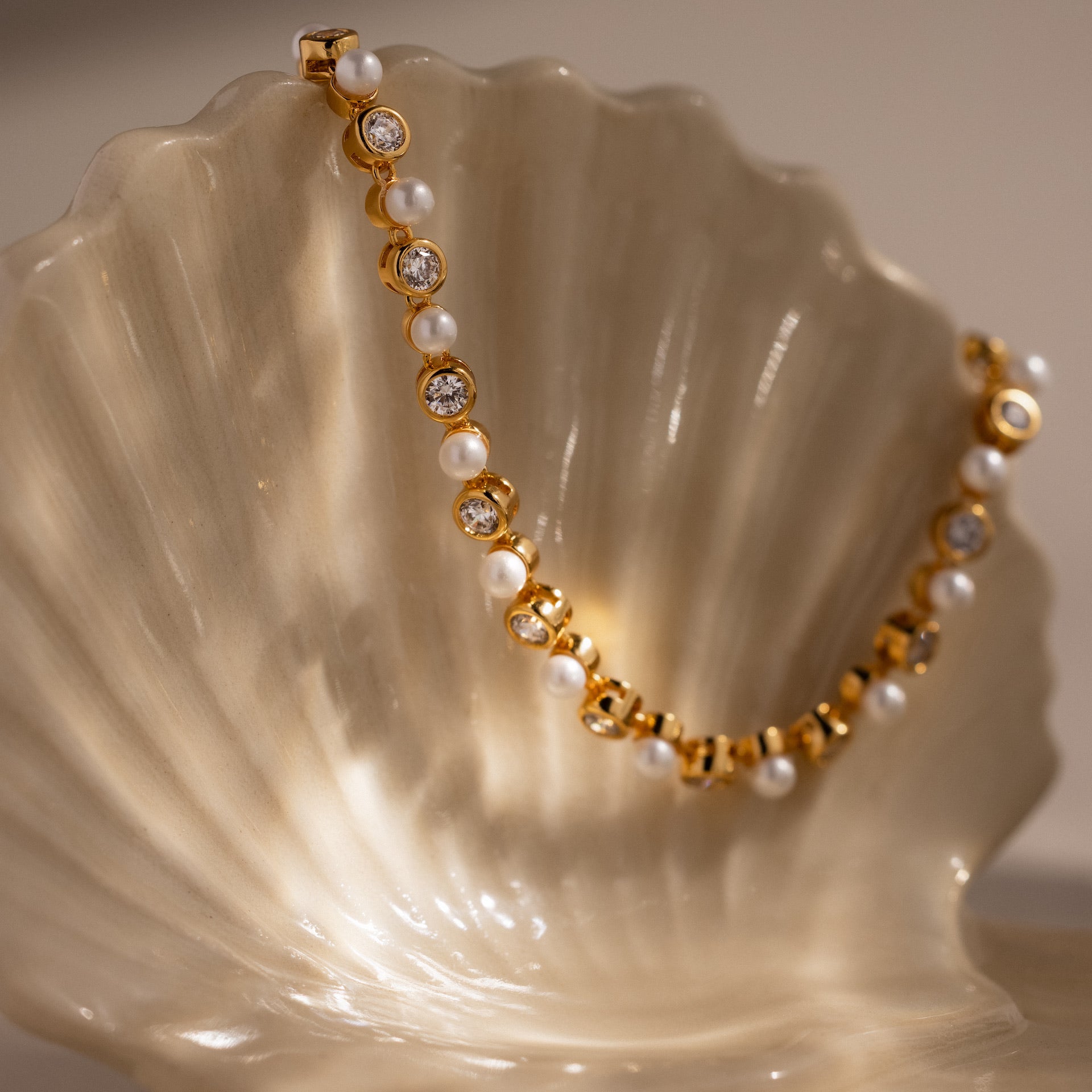 The Pearl & Diamond Tennis Bracelet is draped elegantly over a pearly white seashell, showcasing its luxurious blend of gold, pearls, and diamonds.