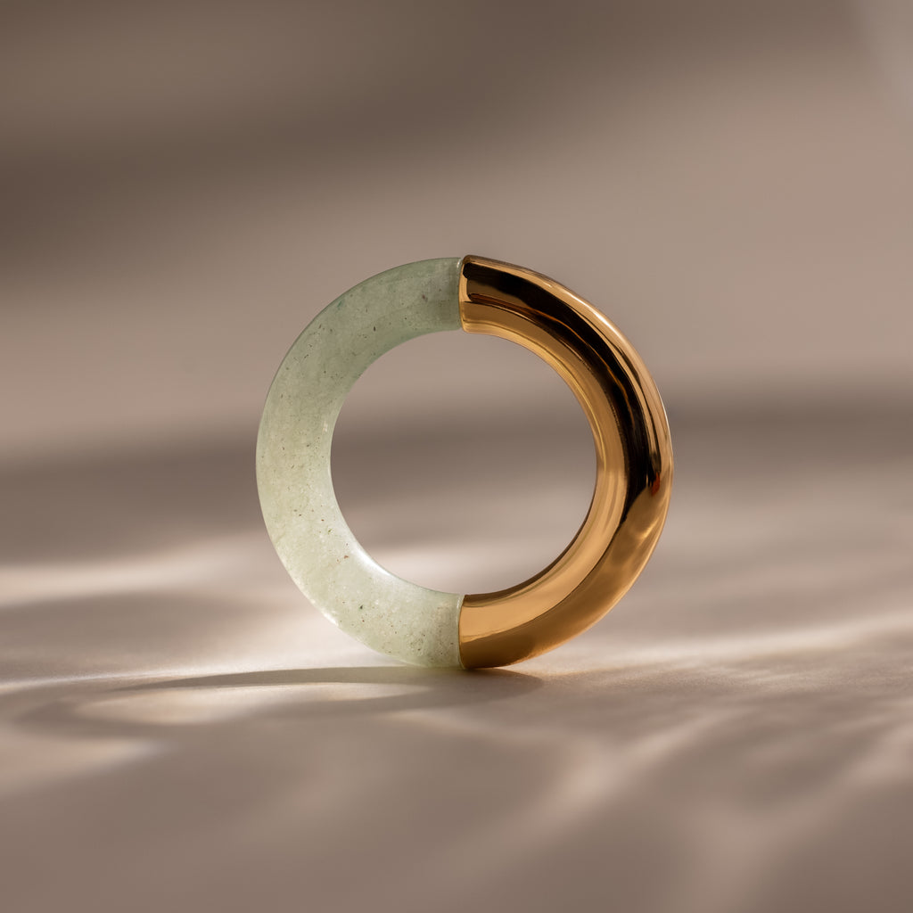 The Half Jade Ring—a circular ring featuring half jade-green stone and half shiny gold—stands upright on a neutral surface, making a striking statement jewelry piece.