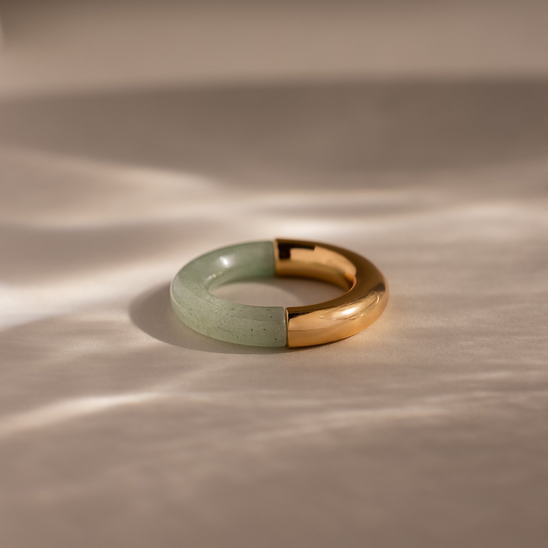 The Half Jade Ring is a round statement jewelry piece with a half-green jade stone and half-gold metal design, displayed on a light, textured surface.