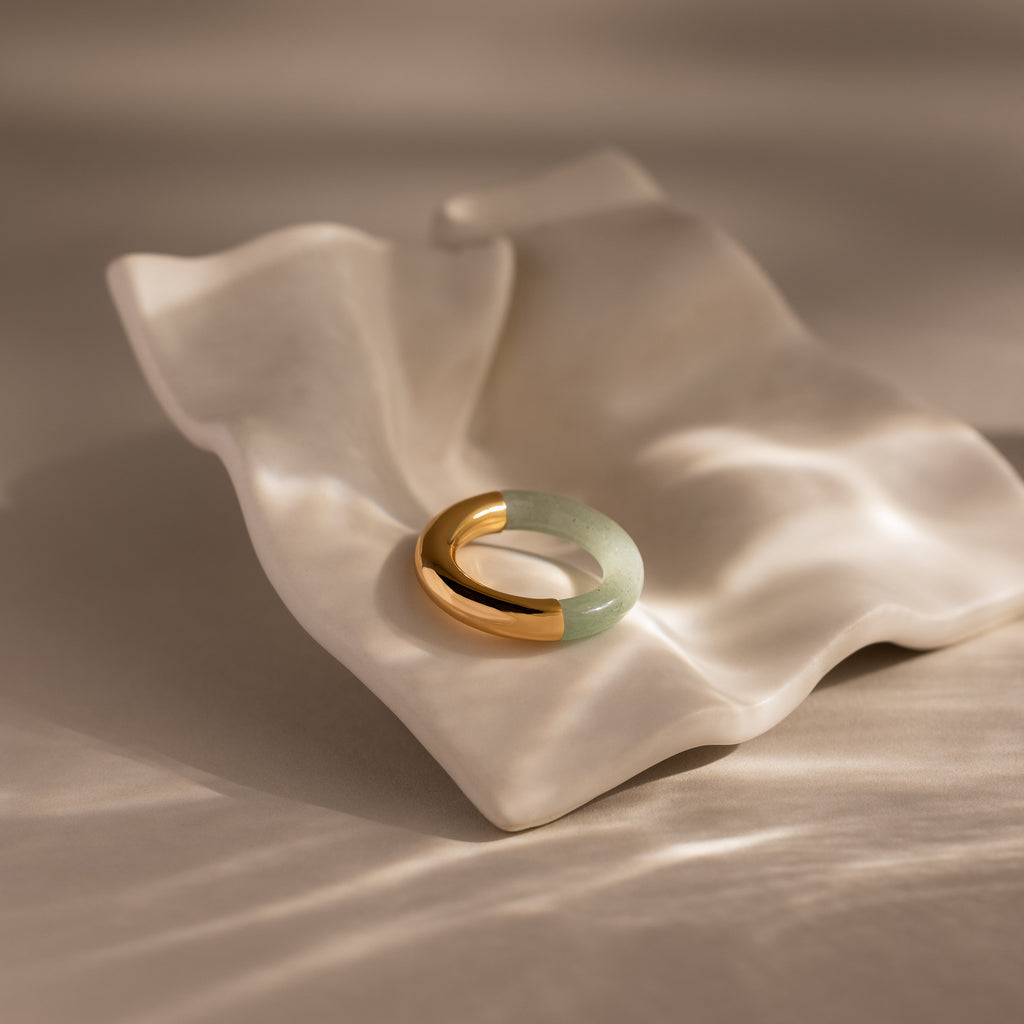 The Half Jade Ring, featuring gold and green jade, rests on a wavy white ceramic dish illuminated by soft natural light.