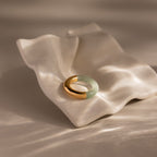 The Half Jade Ring, featuring gold and green jade, rests on a wavy white ceramic dish illuminated by soft natural light.