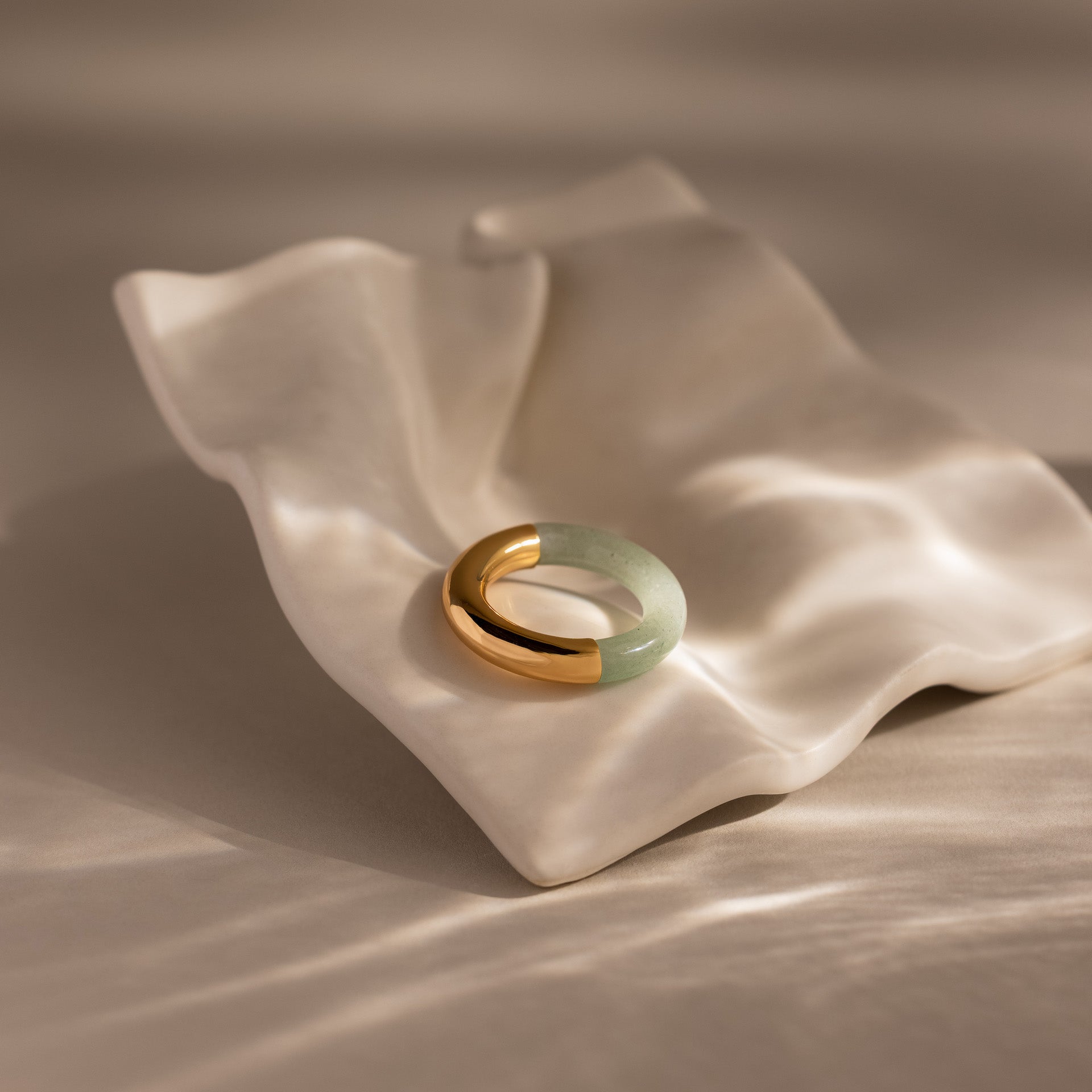 The Half Jade Ring, featuring gold and green jade, rests on a wavy white ceramic dish illuminated by soft natural light.