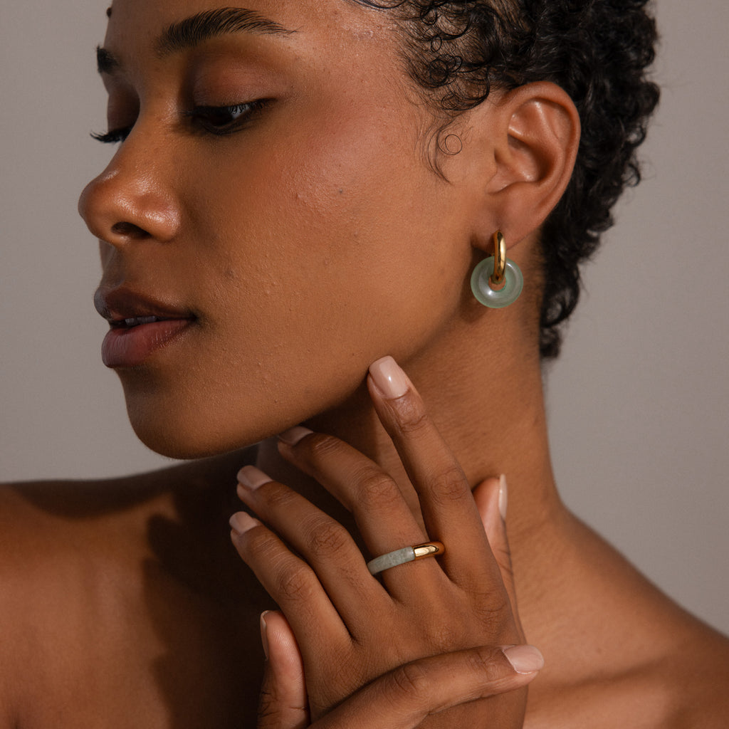 A woman with short curly hair wears the Half Jade Ring and a matching jade hoop earring as she gently touches her neck.